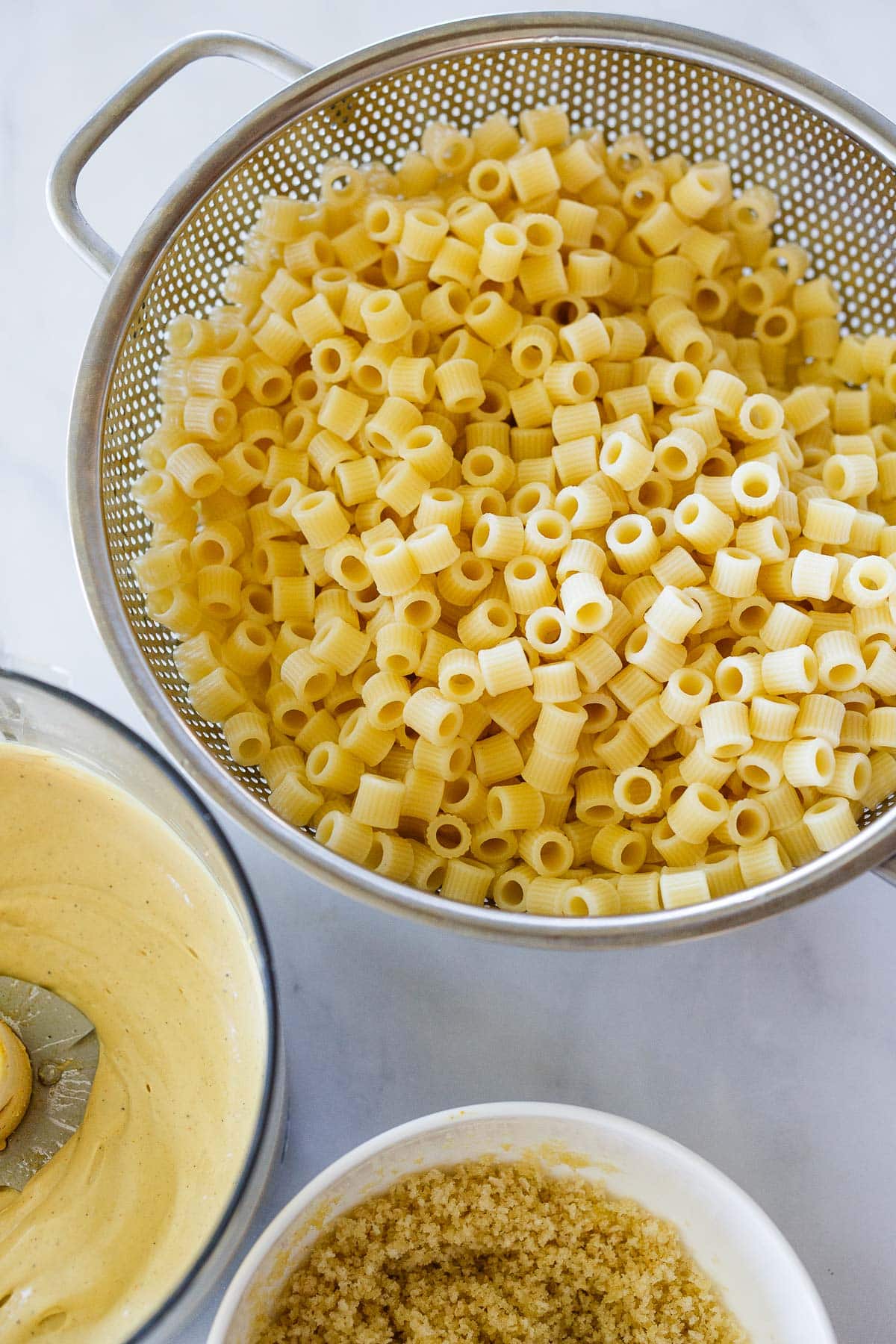 cooked ditalini pasta in a colander next to creamy vegan cheese sauce and bowl of breadcrumb topping.