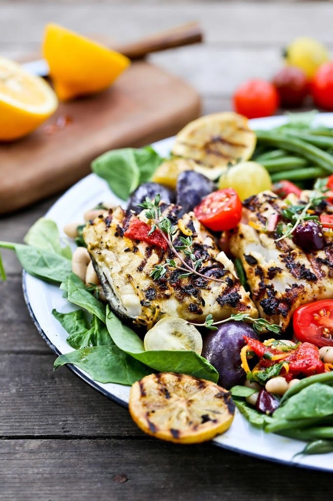 summer nicoise salad with grilled white fish, grilled lemons, tomatoes, white beans, olives, and greens on serving plate.