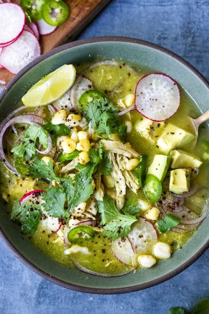 soup bowl with pozole verde, made with chicken, garnished with avocado, jalapeño, radish, red onion, and cilantro, served with lime wedge.