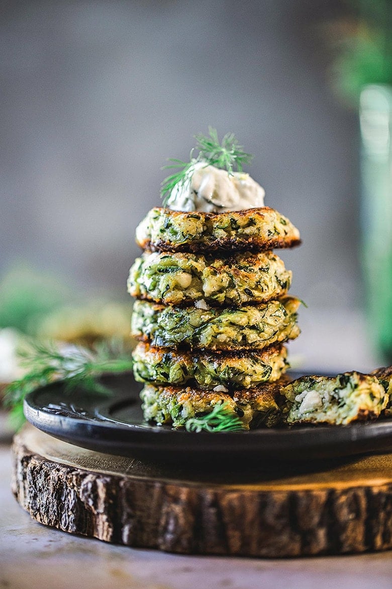 zucchini fritters stacked on plate topped with tzatziki sauce and fresh dill.