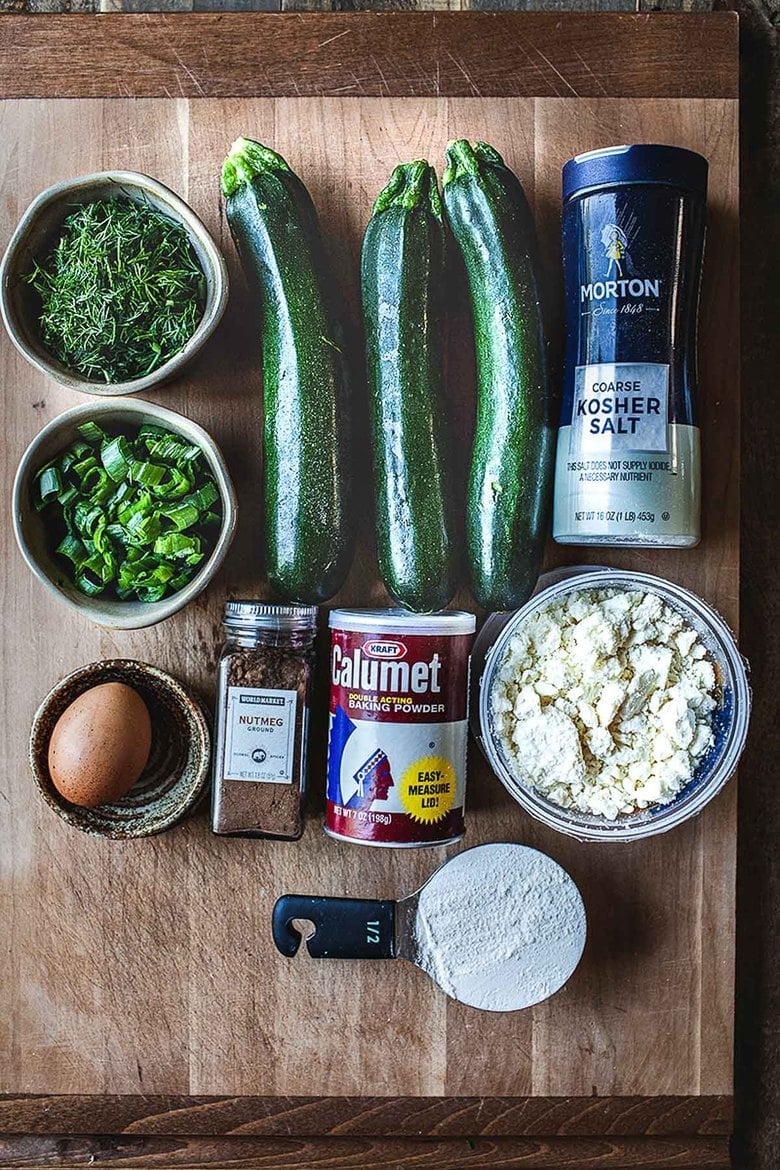 ingredients for zucchini fritters laid out on wood board - kosher salt, zucchini, dill, scallions, egg, nutmeg, baking powder, feta, and flour. 