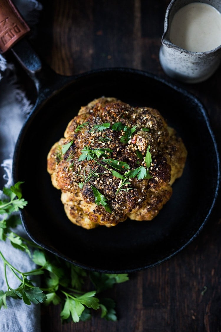 overhead shot of whole roasted cauliflower seasoned with zaatar and garnished with fresh herbs in cast iron skillet.