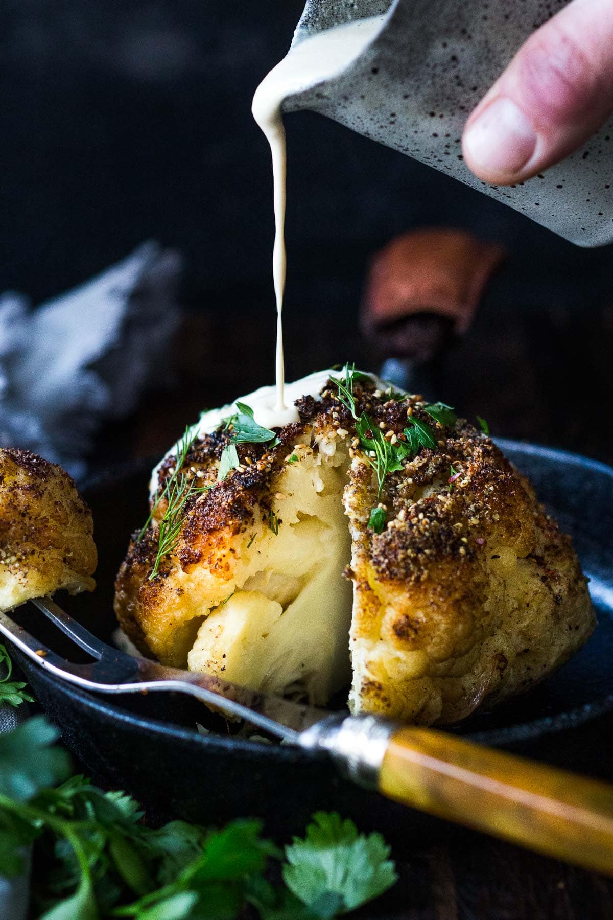  za'taar-spiced, whole-roasted cauliflower with tahini sauce being drizzled over it. 