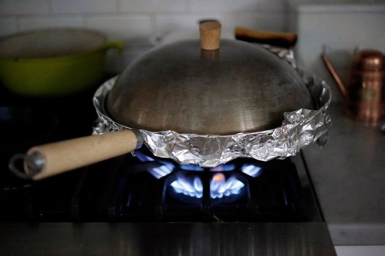 wok covered with foil and lid on a gas burner.
