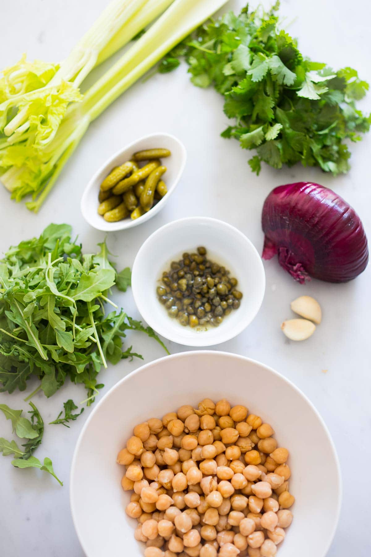 bowls of ingredients laid out for chickpea salad sliders- cornichons, capers, chickpeas, surrounded by celery, parsley, arugula, red onion, and garlic cloves. 