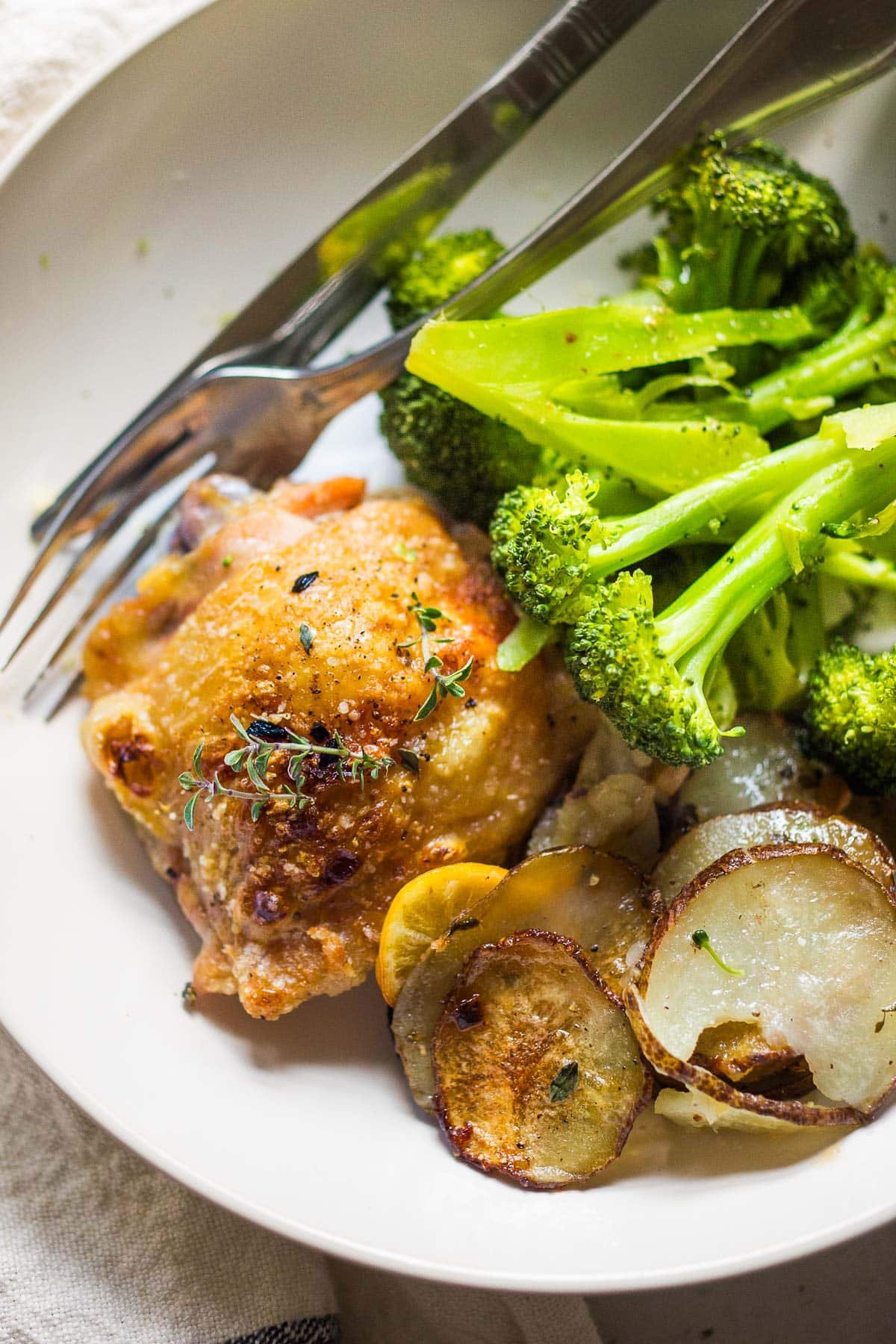 Oven-baked chicken thighs, roasted over thinly sliced potatoes with garlic, lemon and thyme served with broccoli in a white dish. 
