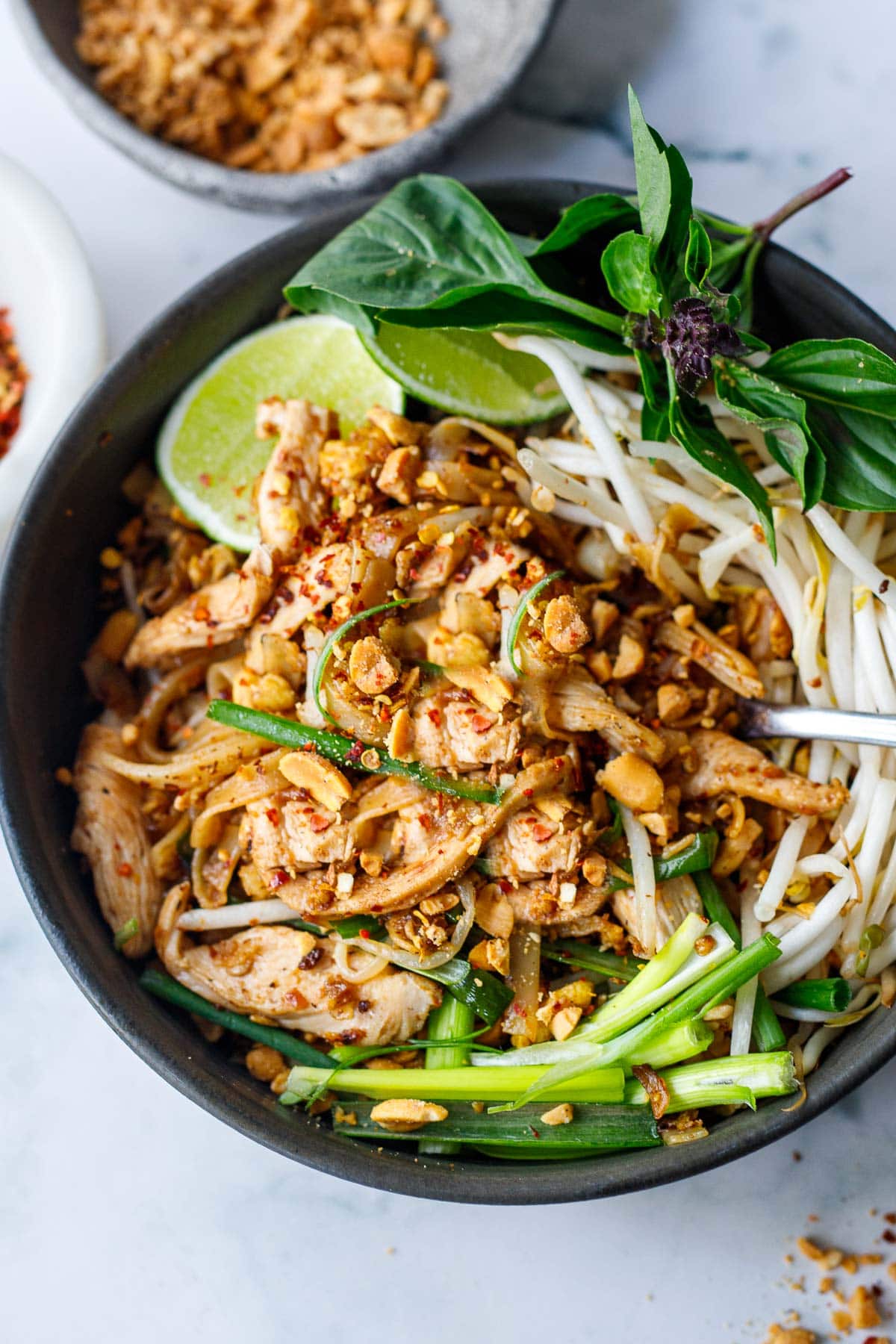A bowl full of Pad Thai with chicken, lime, basil, crush peanuts, bean sprouts and scallions. 