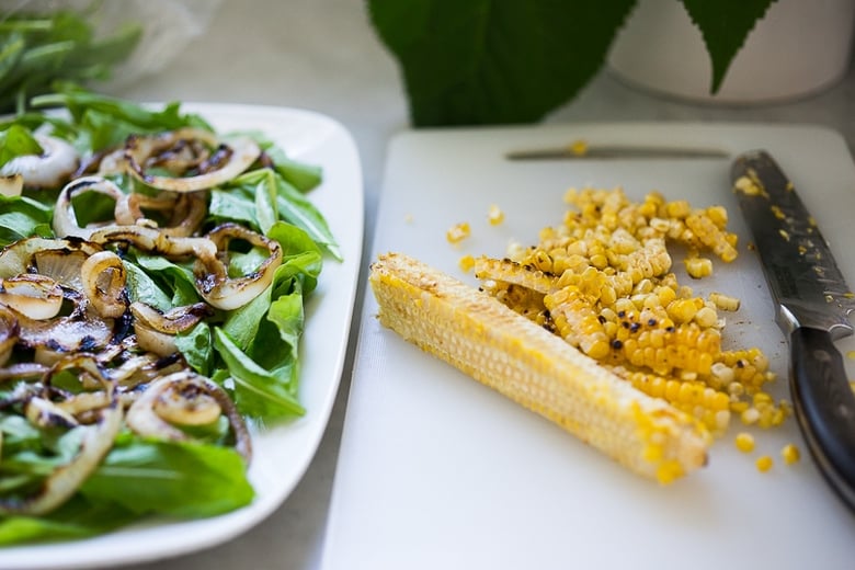 serving platter with greens and grilled onions next to cutting board with sharp knife and grilled corn sliced off the cob.