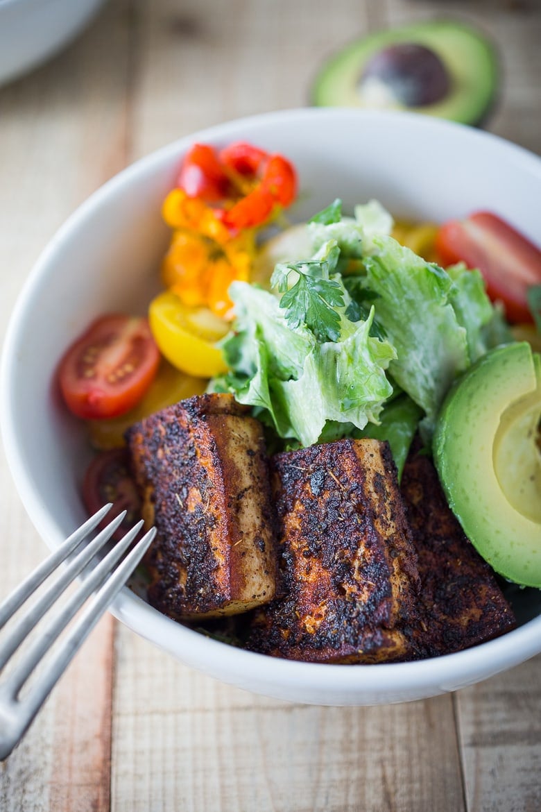 vegan caesar salad with blackened tofu, avocado slices, and cherry tomatoes in bowl.