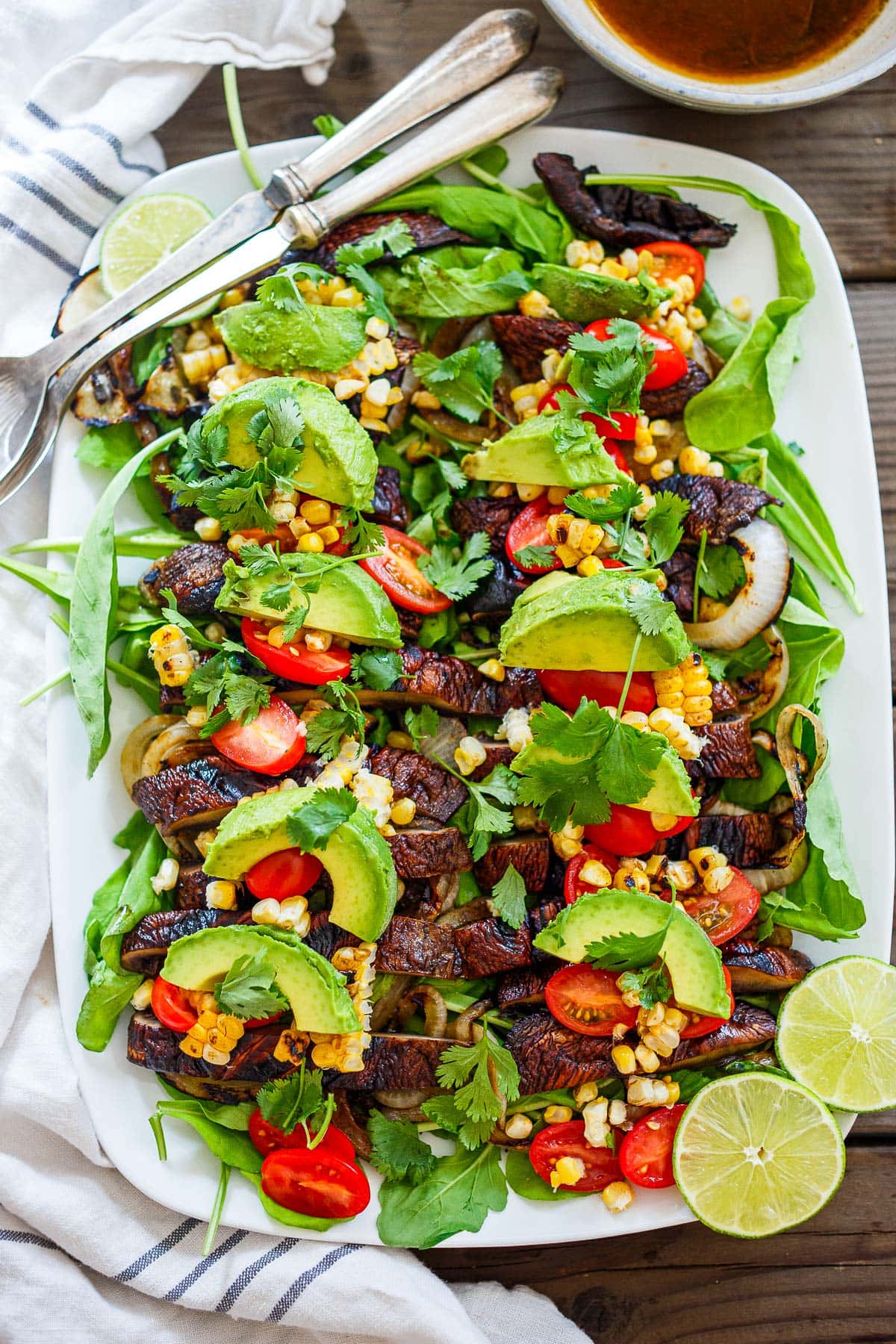 grilled portobello mushroom salad on a white platter with grilled onions and corn, fresh avocado slices and cherry tomatoes, cilantro, and limes.
