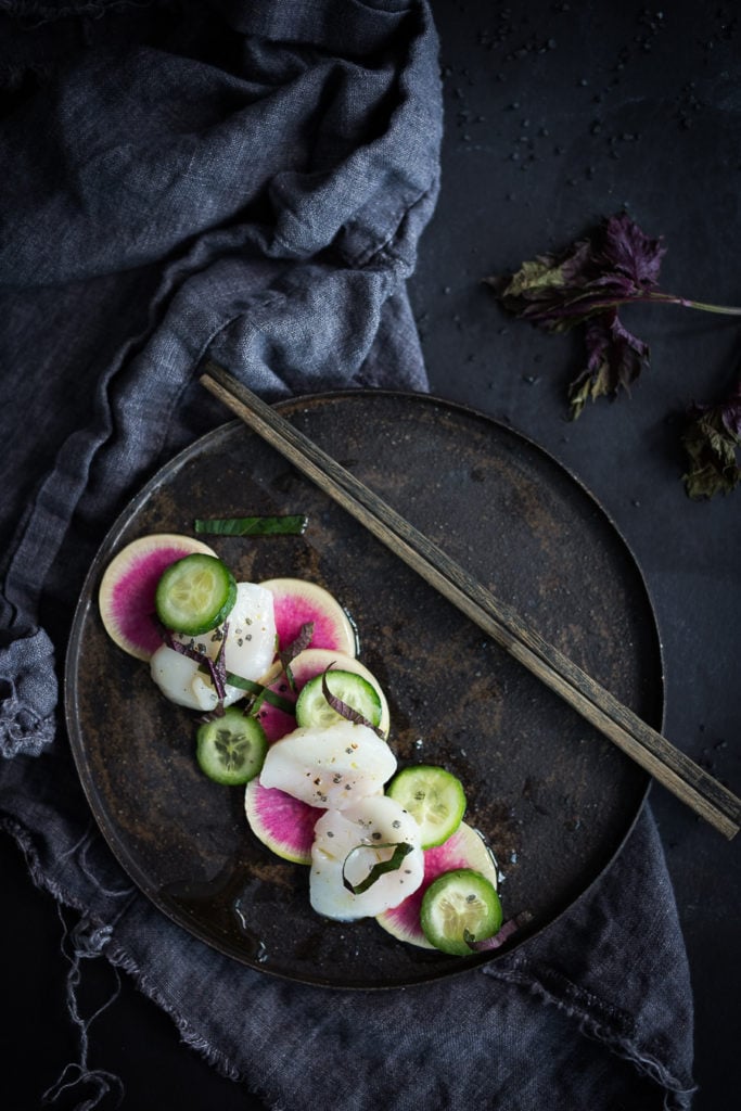 Scallop Crudo on a black plate with chop sticks.
