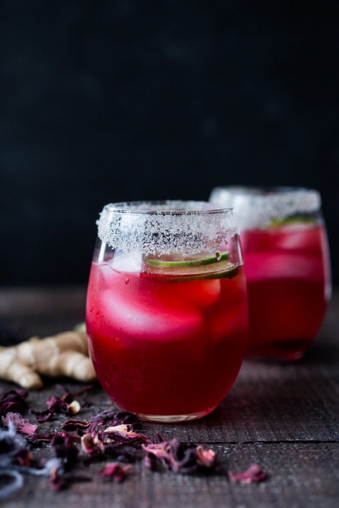 two glasses of hibiscus margaritas with salted rims, topped with lime slice, and fresh ginger beside the glasses.