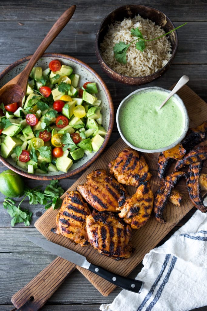 Grilled Peruvian Chicken on a board with a bowl of Cilantro Sauce and Avocado Tomato Salad with a bowl of rice.