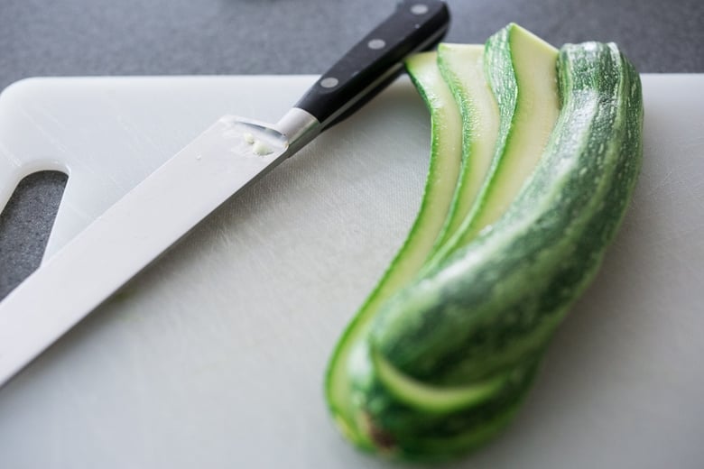 cutting board and sharp knife with zucchini sliced thinly in long strips. 