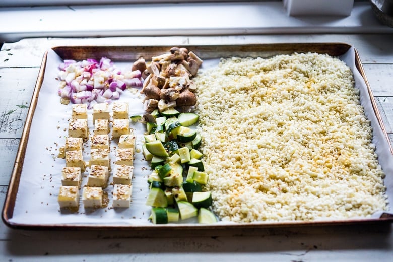 baking sheet with parchment paper with riced cauliflower spread out, diced mushrooms and zucchini, chopped red onion, and cubed tofu arranged in sections.