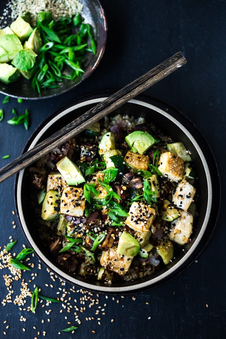 cauliflower rice bowl topped with roasted mushrooms and zucchini, baked sesame tofu, and green onions with chopsticks laying on top of the bowl.