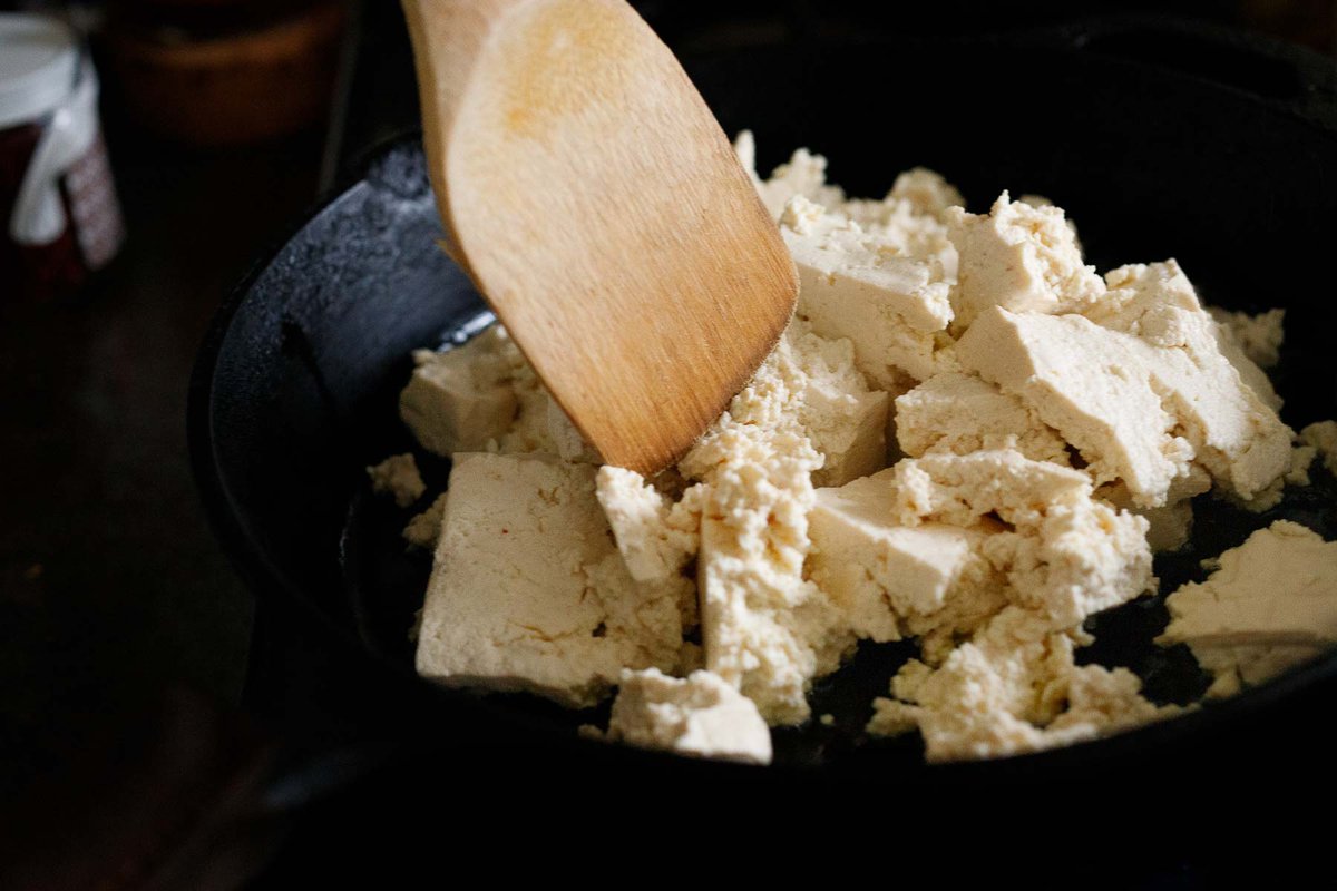 breaking apart tofu in a skillet with a wood spatula.