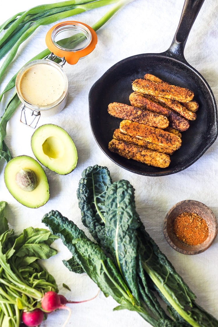 ingredients for blackened tempeh salad laid out - blackened tempeh in cast iron, cajun ranch dressing, avocado, cajun spice, lacinato kale, radishes.