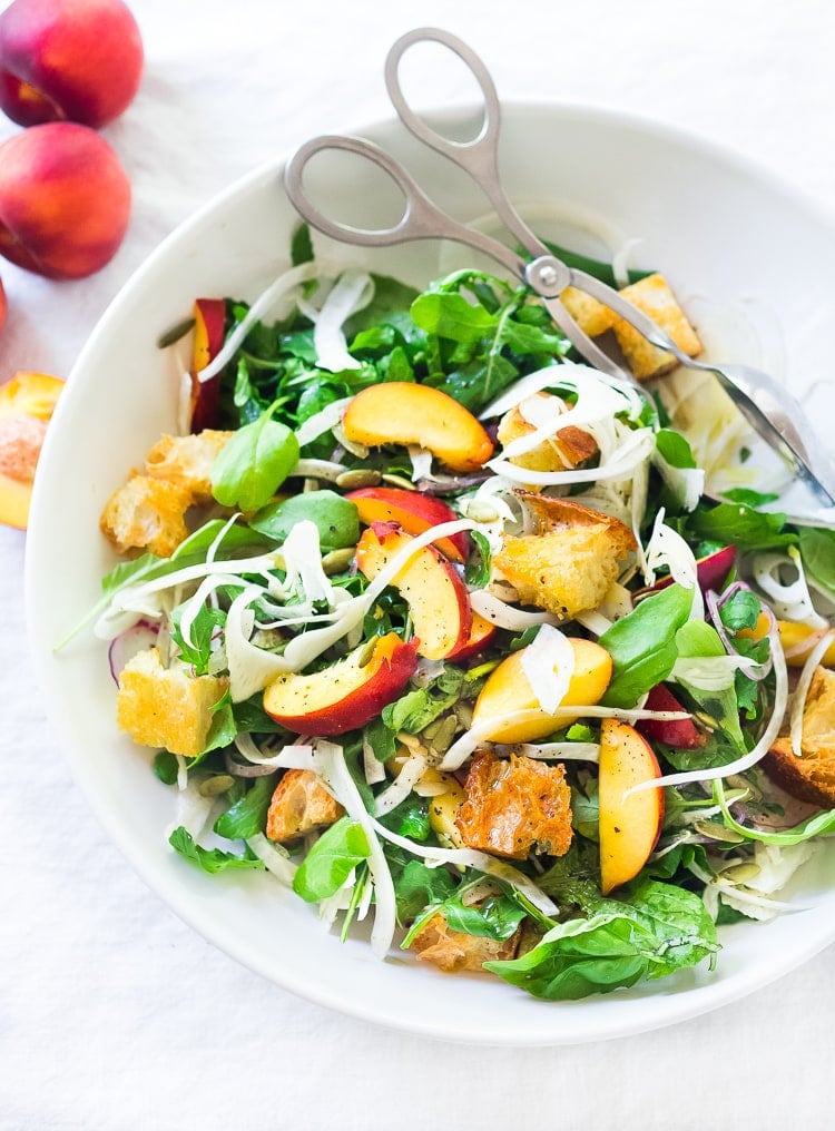 salad bowl with salad tongs with peach panzanella salad topped with fennel and served with arugula.