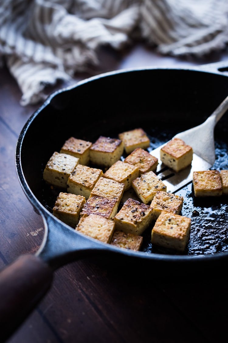 cast iron skillet with metal spatula lifting cubes of seasoned crispy tofu.