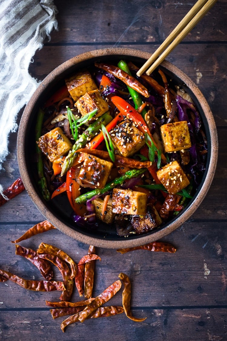 bowl of szechuan tofu and veggies with chopsticks, garnished with sesame seeds and green onions - dried chilies next to the bowl.