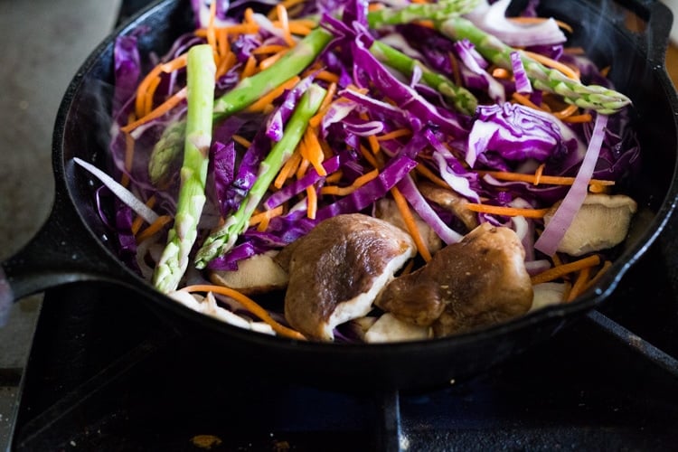 sliced red cabbage and red onion, mushrooms, asparagus, and shredded carrots in cast iron skillet cooking.
