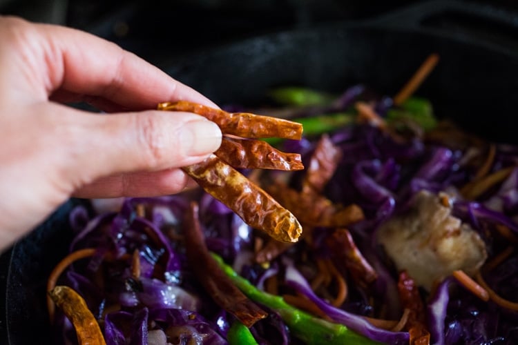 hand holding three dried red chilies over skillet with sautéed veggies.