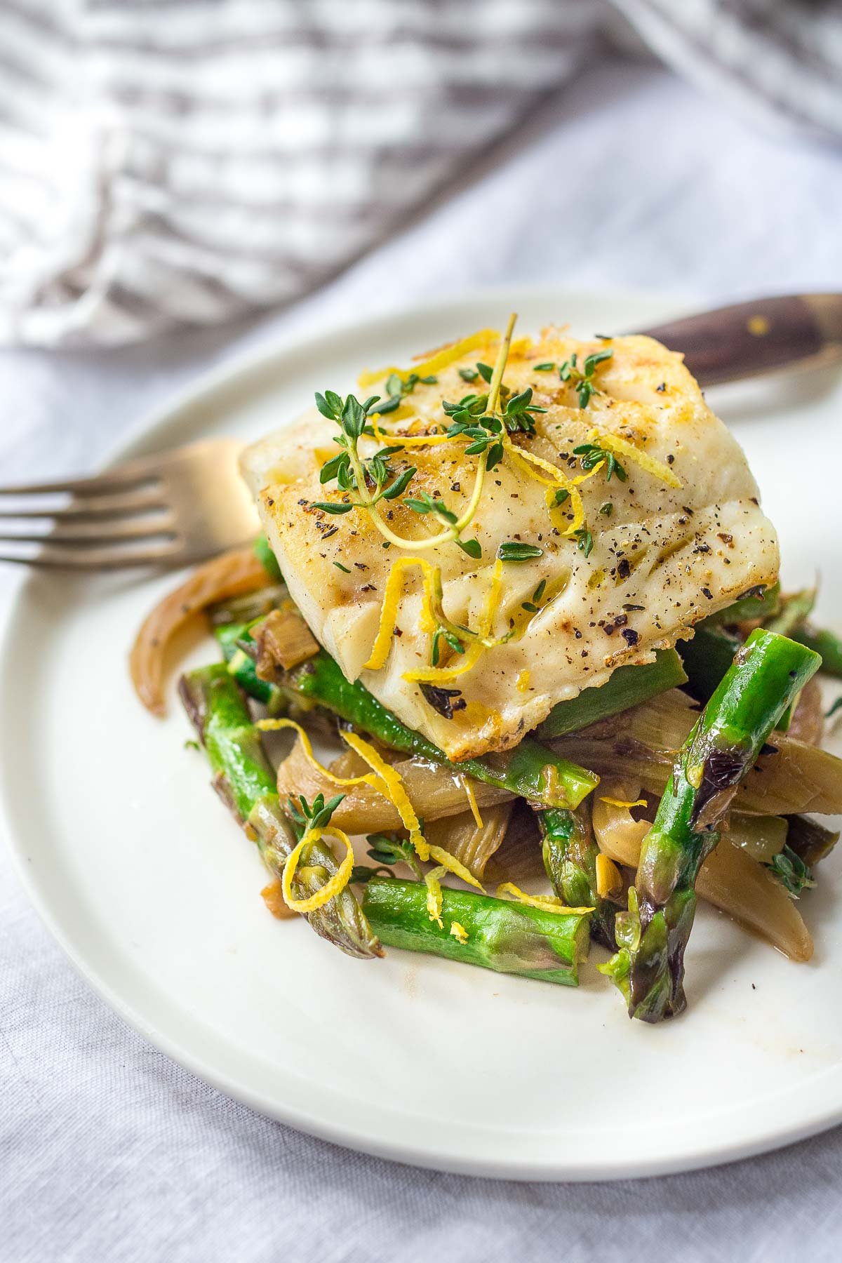  Baked cod with lemon zest and thyme served over spring veggies on a white plate. 