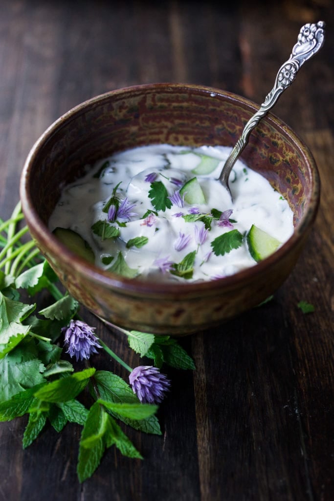 Raita in a brown pottery bowl with chive blossoms and cilantro.