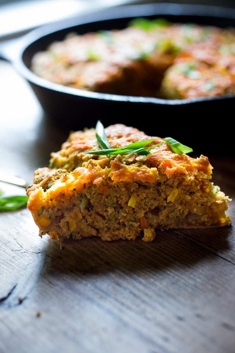 slice of zucchini cornbread with cheddar topping and green onion garnish, in front of cast iron skillet.