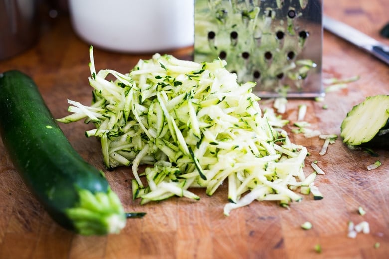 grated zucchini on wood cutting board next to zucchini and grater.