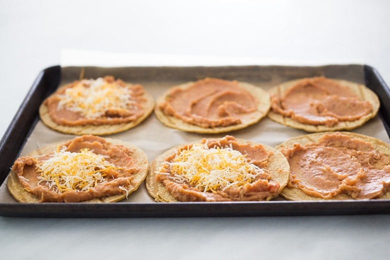 six tortillas on baking sheet with refried beans spread over top and three tacos with shredded cheese.