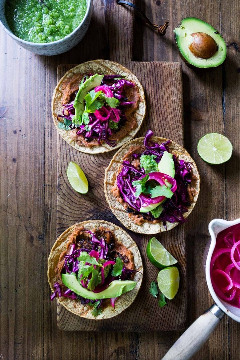 wood serving board with three jackfruit tacos topped with cabbage slaw, salsa, avocado, pickled onions, and cilantro, served with lime wedges.