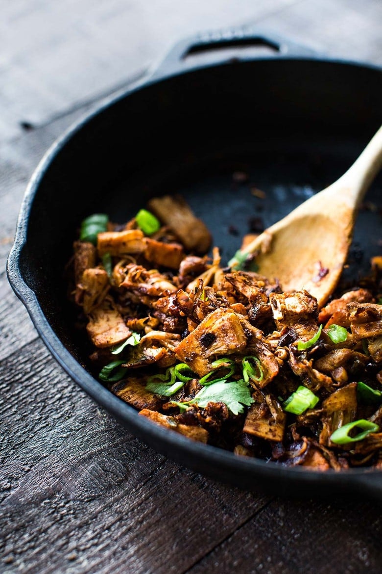 wood spoon stirring crispy jackfruit carnitas in cast iron skillet with green onions and cilantro.
