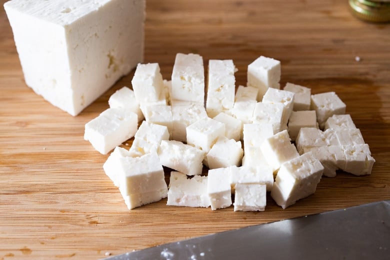 wood cutting board and sharp knife with feta block and cubed feta.
