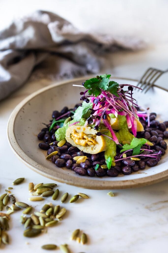 butternut squash tamale on a bed of black beans, topped with pumpkin seed cilantro sauce, watermelon radishes, microgreens, and fresh cilantro.