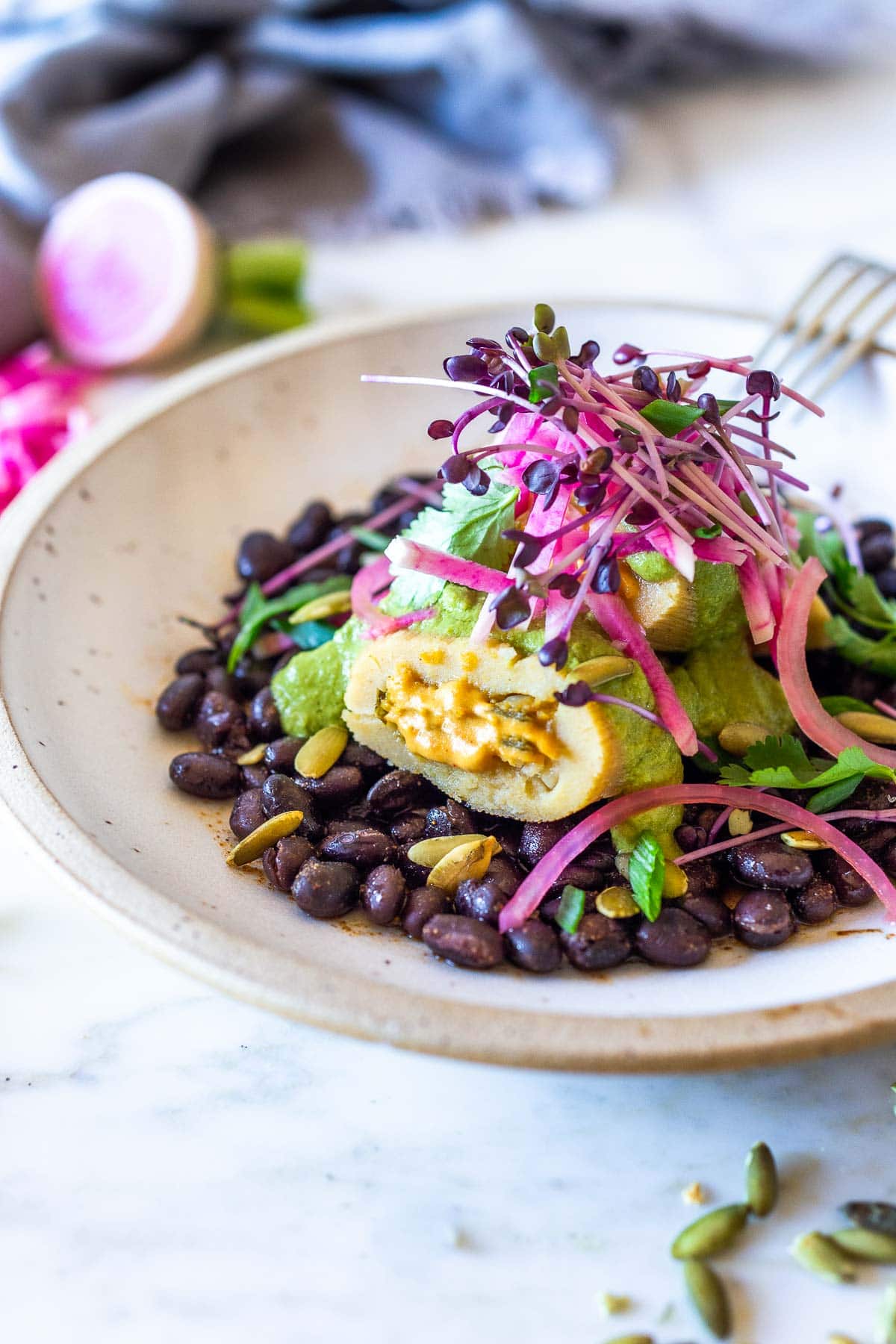 Vegetarian tamales in a white bowl with black beans, poblano sauce, watermelon radishes, cilantro and and microgreens  