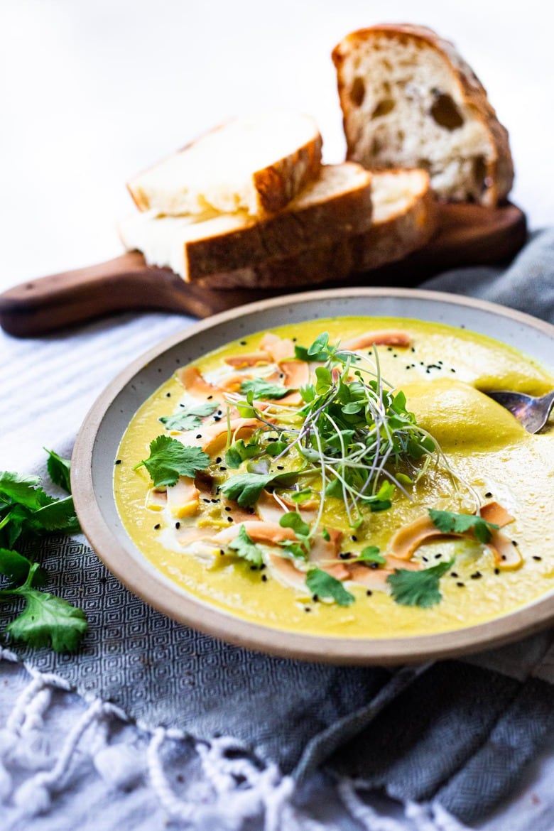 bowl of curried cauliflower soup garnished with toasted coconut, nigella seeds, cilantro, and micro greens next to wood board with sliced bread. 