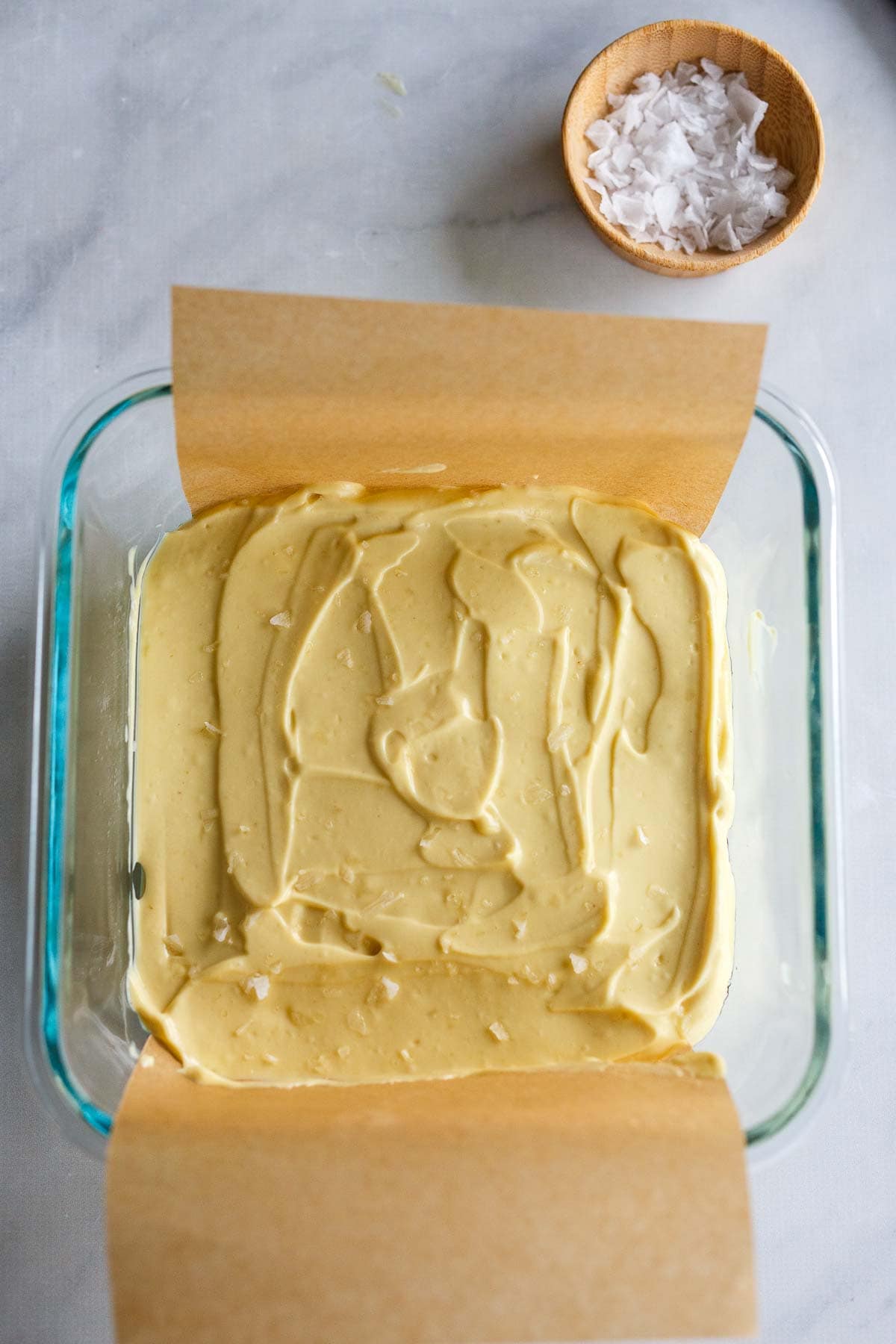 creamy vegan parmesan cheese mixture in glass dish with parchment paper with a smooth surface and coarse salt sprinkled over top.
