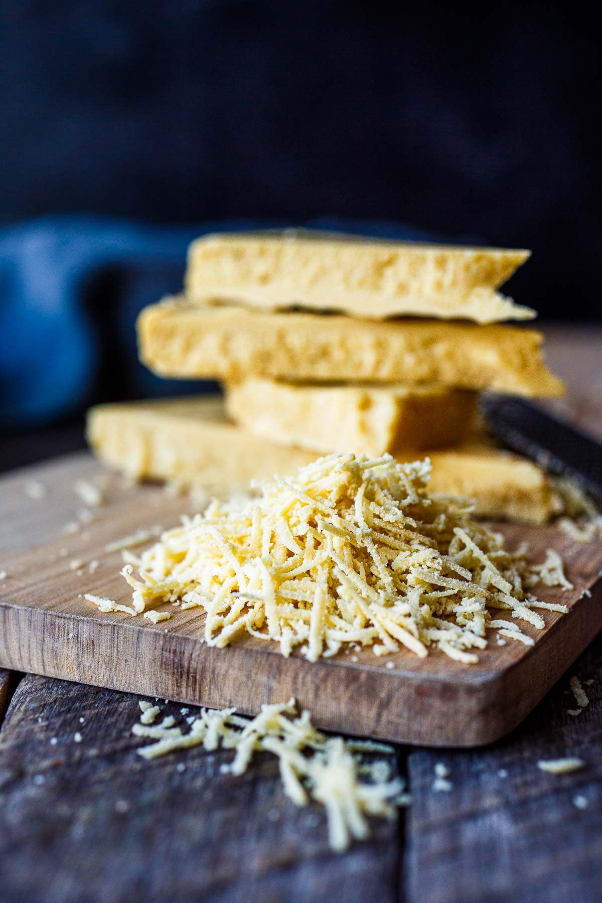 grated homemade vegan parmesan on wood cutting board with wedges of cheese stacked in background.