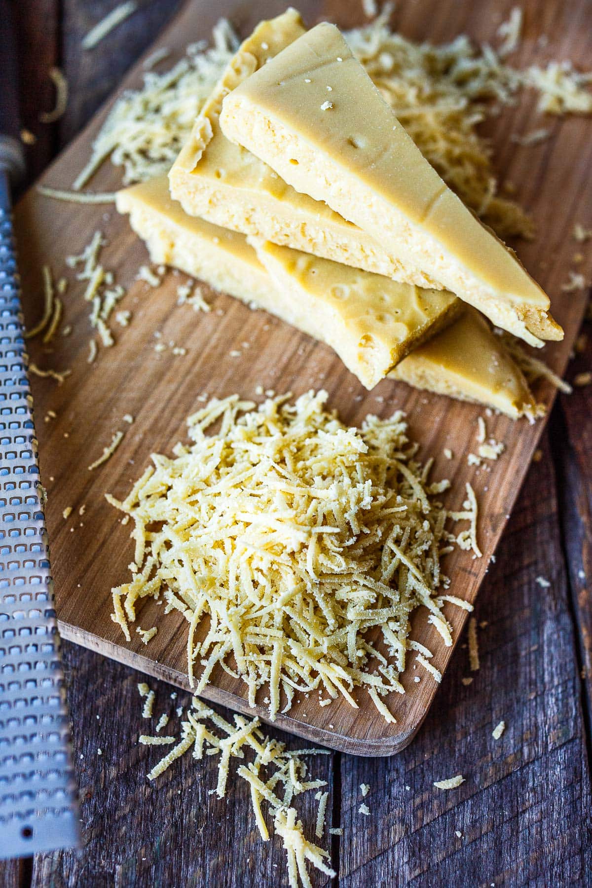 wood cutting board with a pile of grated vegan parmesan cheese and a stack of parmesan wedges.