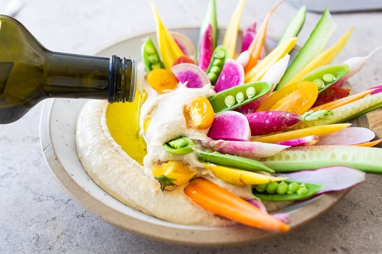 drizzling olive oil onto hummus with rainbow veggies tucked into dip.