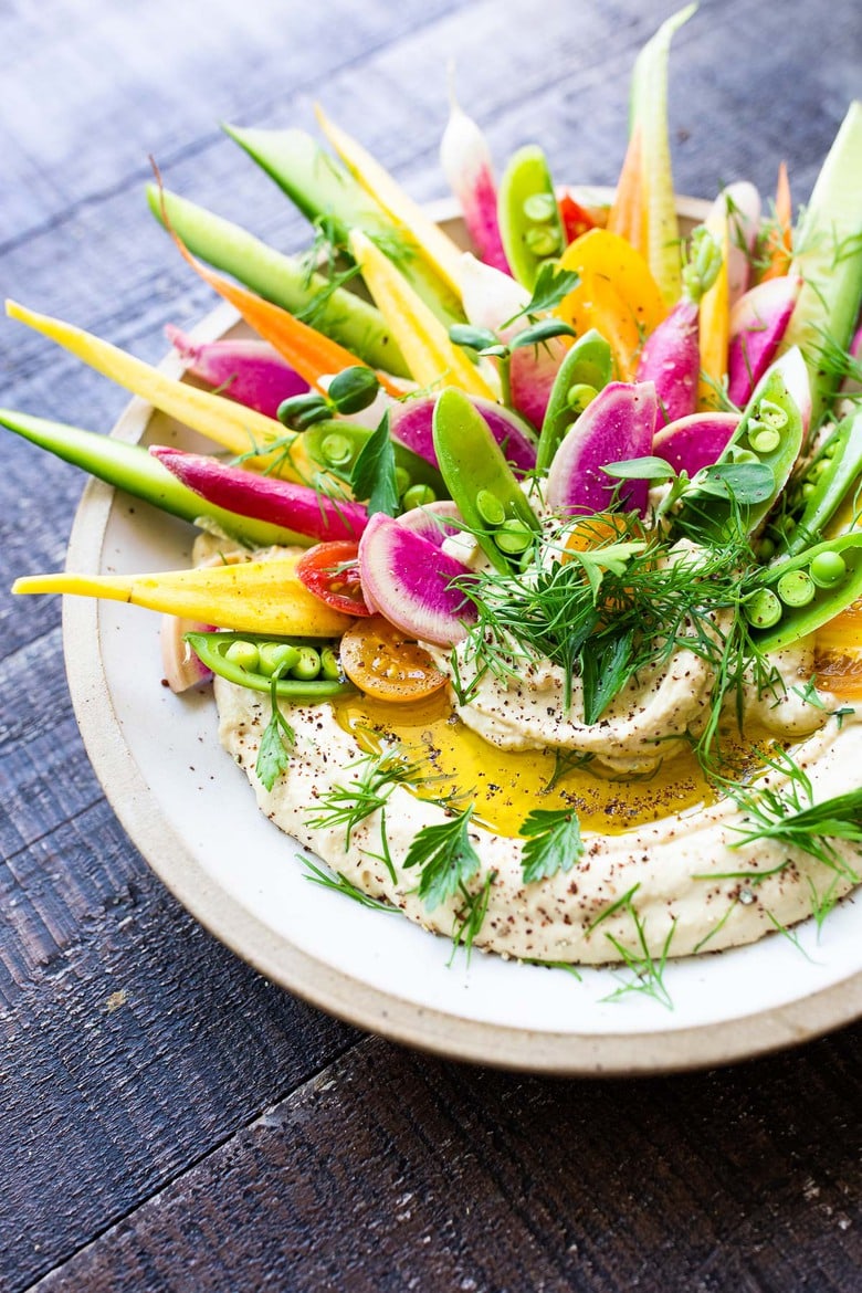 hummus, garnished with olive oil, fresh herbs, and rainbow veggies, sprinkled with spices.
