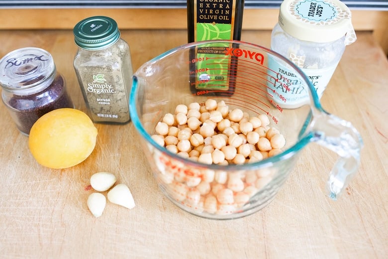 hummus ingredients on wood surface - measuring cups with chickpeas, garlic cloves, lemon, sumac, cumin, olive oil, tahini.