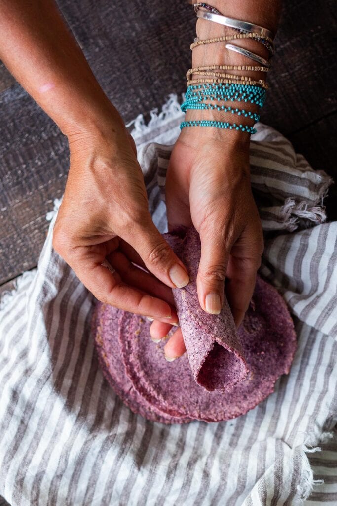 hands rolling blue corn tortillas over pile of homemade tortillas. 