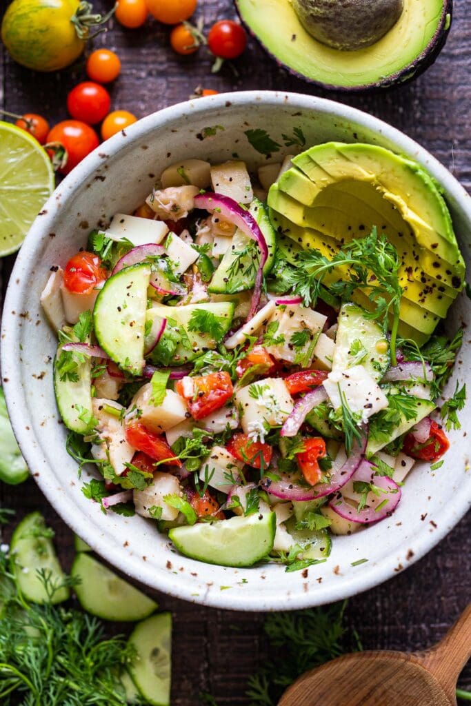 hearts of palm ceviche in bowl with pickled onion, sliced cucumber, sliced avocado, and fresh herbs.