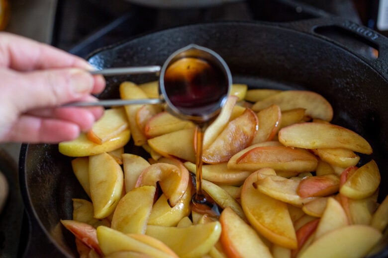 pouring maple syrup into cast iron skillet with softened apples. 