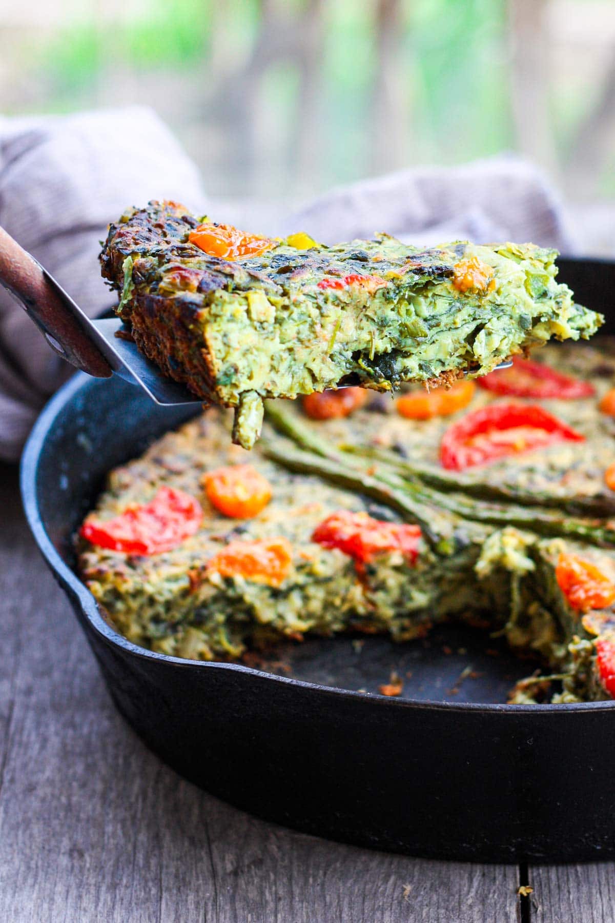 cast iron pan with green veggie vegan frittata, topped with tomatoes, spatula holding slice over pan.