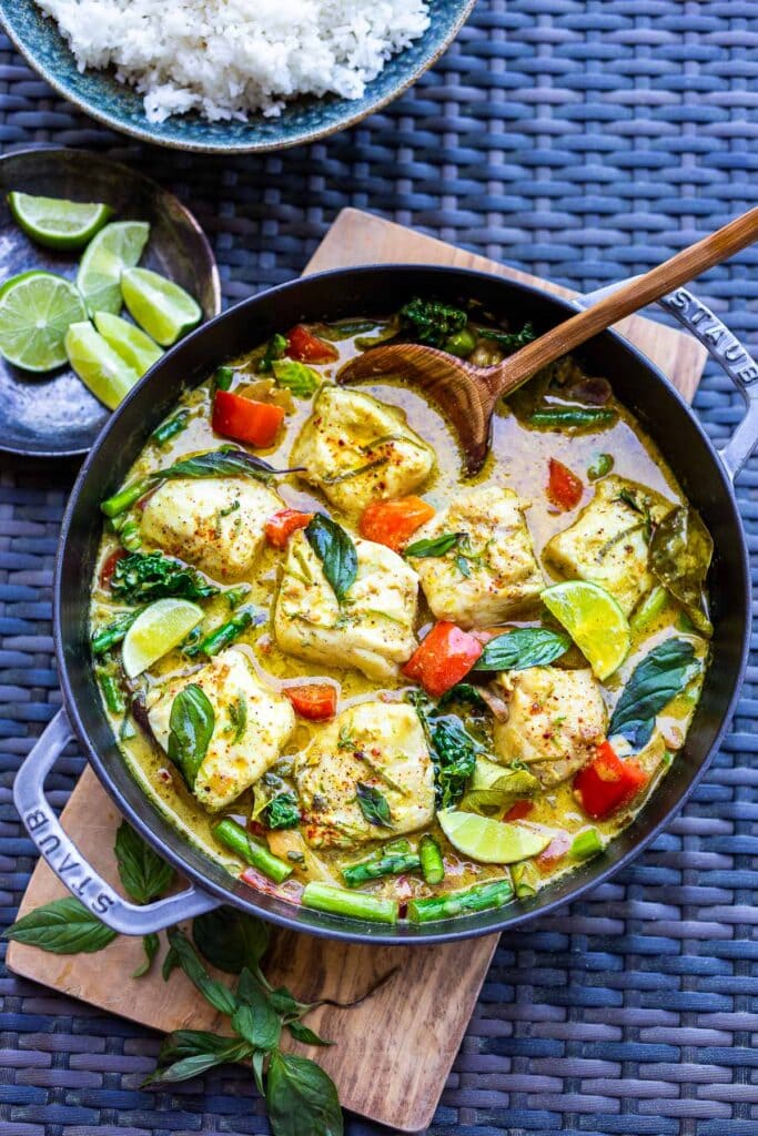 dutch oven with thai fish curry and wooden spoon next to small plate of lime wedges and bowl of rice.