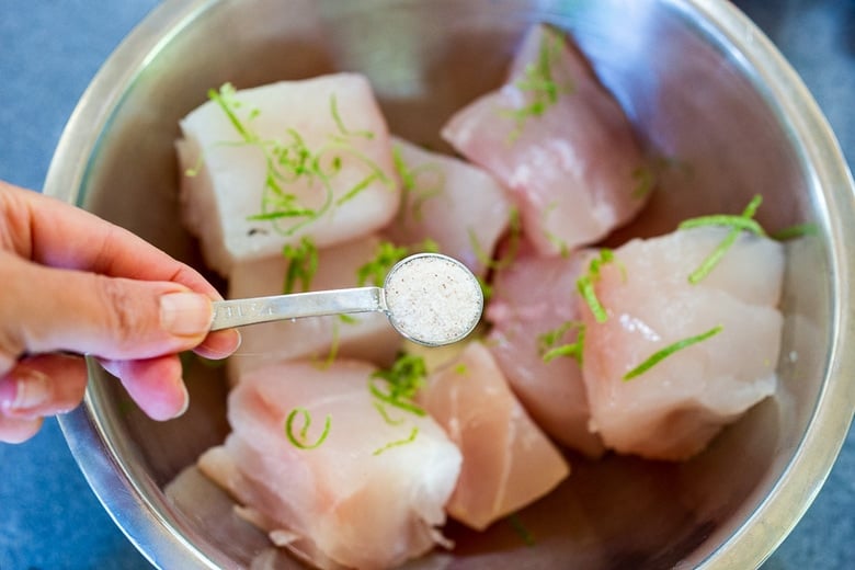 mixing bowl with white fish and lime zest, teaspoon of salt held over top.